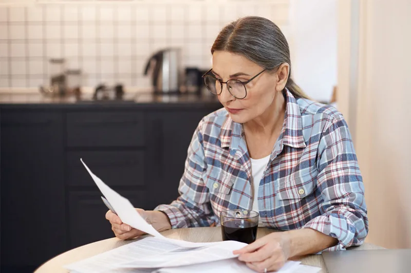 Woman wearing glasses with a focused expression sitting at a kitchen table reading paperwork with a coffee cup, symbolising personal reflection and navigating post-relationship decisions.