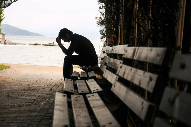 Man sitting alone on a bench under a shelter, the beach in the distance, leaning forward with his head resting in his hands, conveying grief and contemplation.