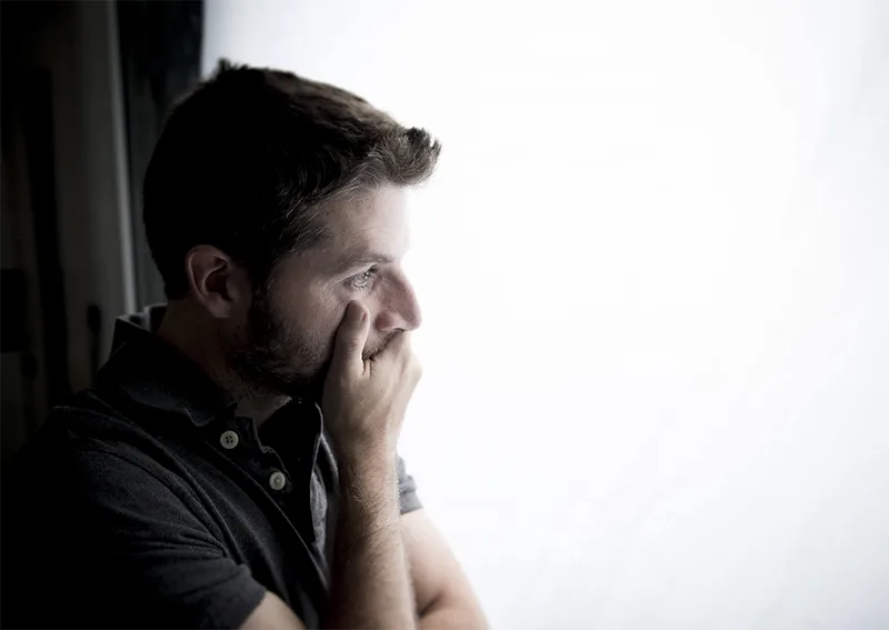 Man standing at a window with one hand covering his mouth and the other arm across his chest, symbolising the emotional weight and complexity of separation.