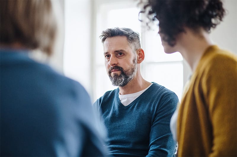 Couple in a YMU relationship counselling intensive session, woman looking attentively at her male partner while the therapist listens with their back to the viewer.