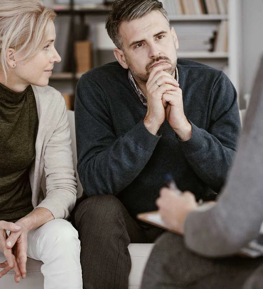 Couple sitting on a couch in session with a therapist; woman looks at her partner with care while he listens thoughtfully with hands clasped under his chin.