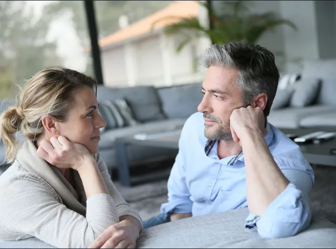 Couple sitting at a table, resting their heads on their hands and sharing soft smiles, representing hope and connection during relationship challenges.