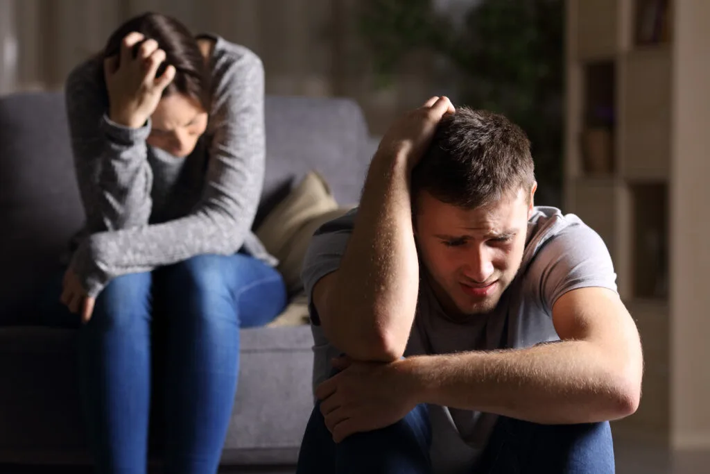 Distressed couple mirroring each other’s posture; woman on couch and man on floor both cradling their heads and wrapping arms around themselves, symbolising shared emotional pain and disconnection.