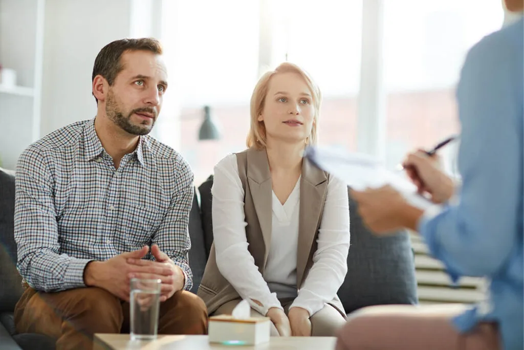 Couple sitting close but not touching on a therapist’s couch, both intently focused on the therapist who is taking notes—illustrating the reflective nature of discernment counselling sessions.