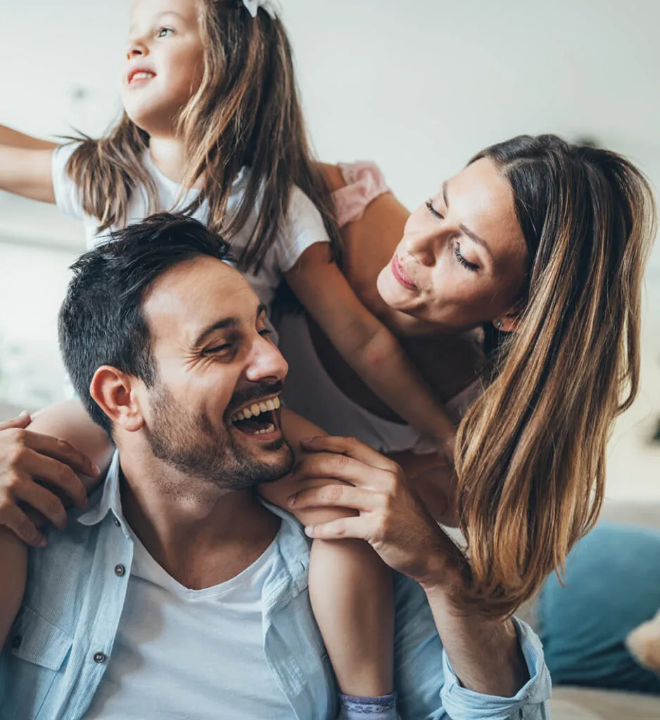 Smiling young family with daughter on father’s shoulders and mother embracing them both, symbolising connection, joy, and restored relationships.