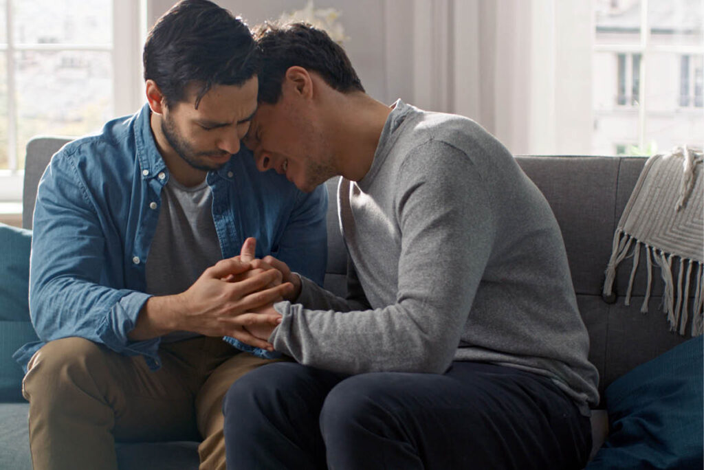 Same-sex male couple sitting closely on a couch, one resting his head gently on the other’s shoulder, hands clasped, expressing comfort and emotional support during relationship counselling.