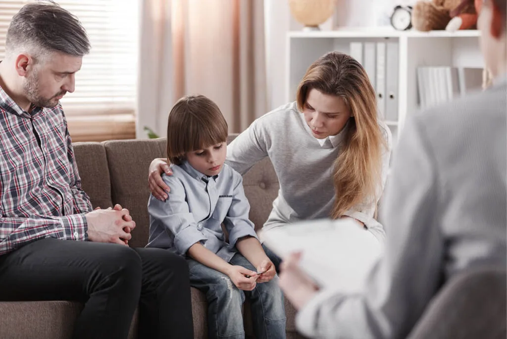 Couple and their young son sitting on a therapist’s couch; mother with arm around son, who is focused on an object in his hands, father watching him closely—depicting a family in emotional reflection during discernment counselling.
