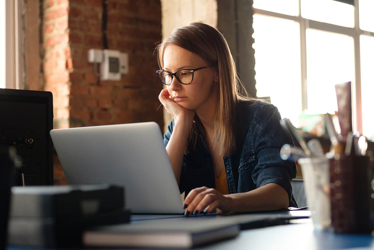 Young woman in an office setting working on her laptop, resting her head in one hand, reflecting the emotional weight of solo private practice