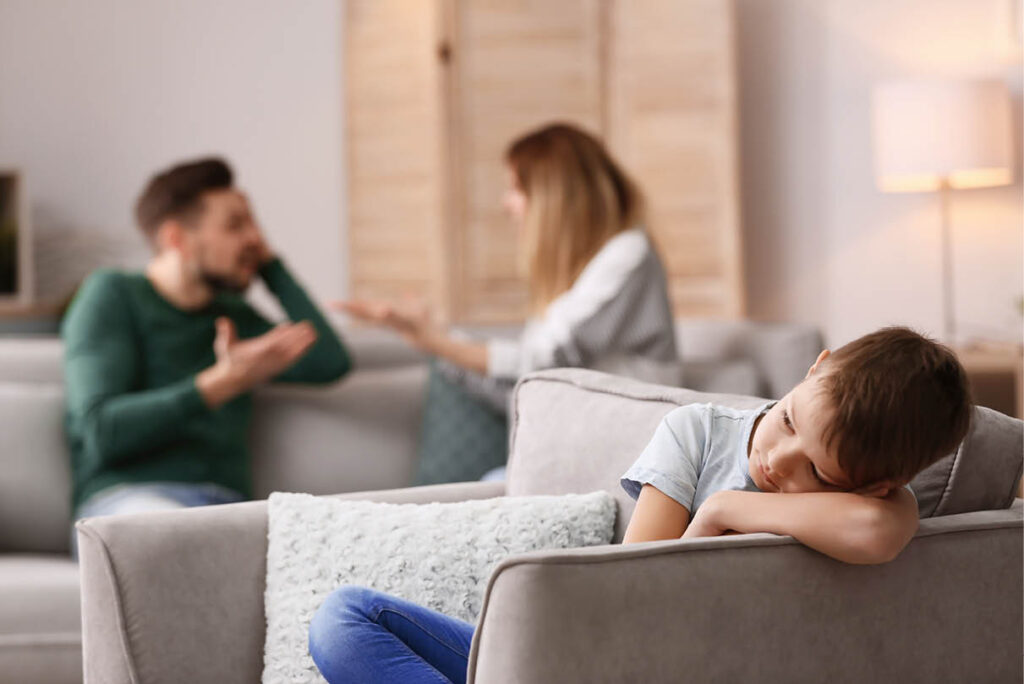 Young boy sitting alone with his head resting on his arm, turned away from his arguing parents in the background, illustrating the emotional toll of family conflict
