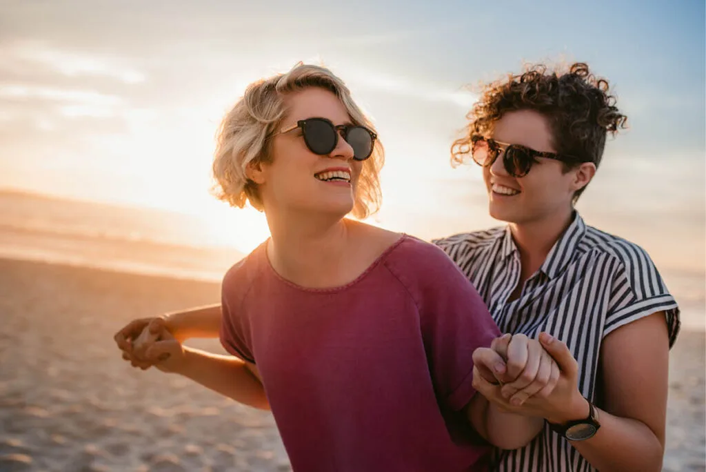 Same-sex female couple smiling and holding hands, one in front of the other, on a sunlit beach, symbolising love, connection, and relationship healing.