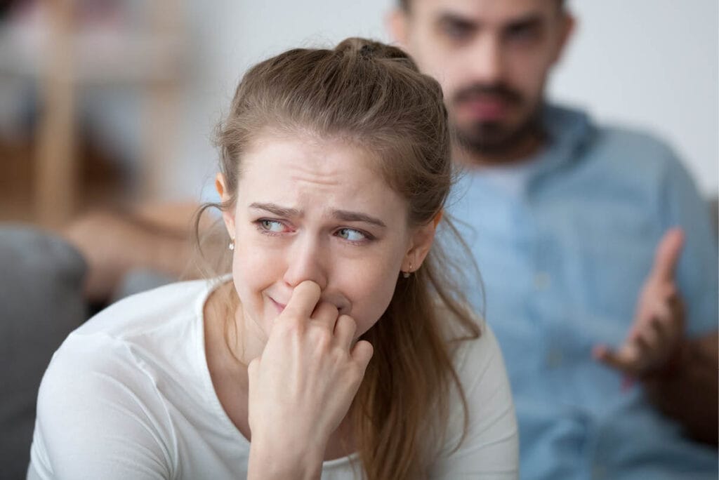 Woman visibly distressed, holding her hand to her mouth, while her partner gestures in frustration in the background, reflecting communication breakdown during an argument