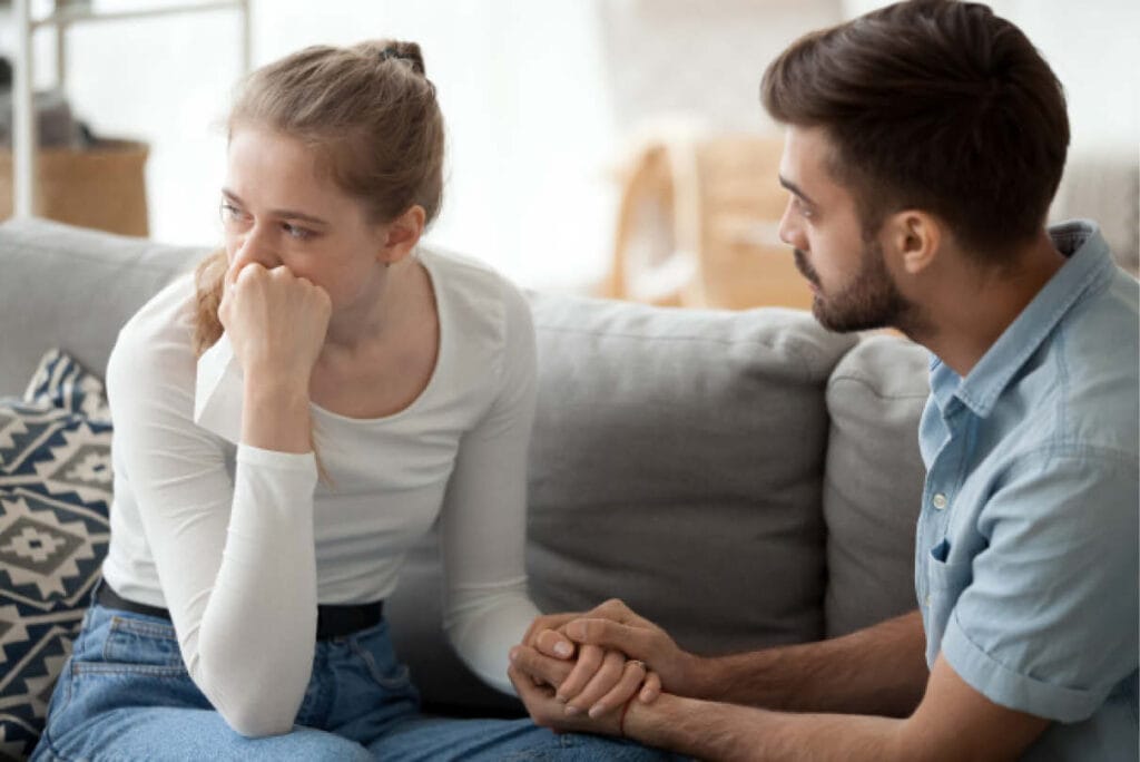 Couple sitting on a couch during therapy; man gently clasping both hands around his partner’s hand while she looks away, holding a tissue, conveying emotional distance and a desire to reconnect.
