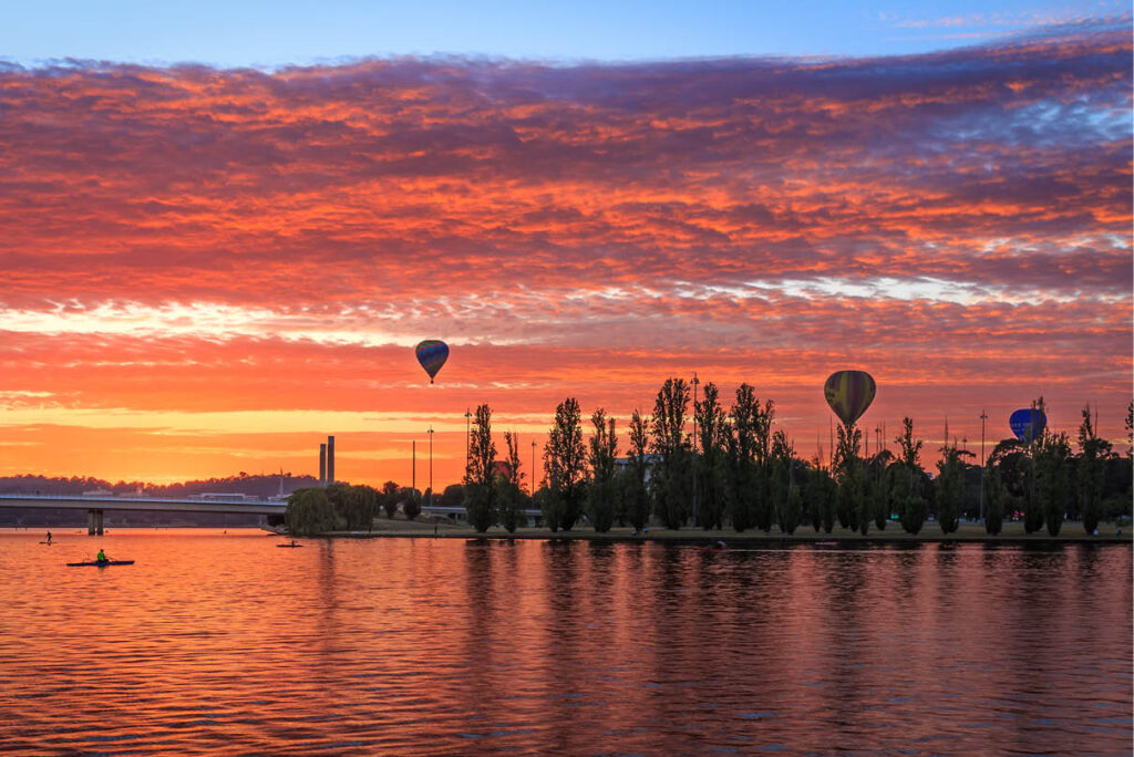 Sunrise over Lake Burley Griffin in Canberra, featuring vibrant reds, yellows, and purple skies with colourful hot air balloons drifting peacefully above the water.