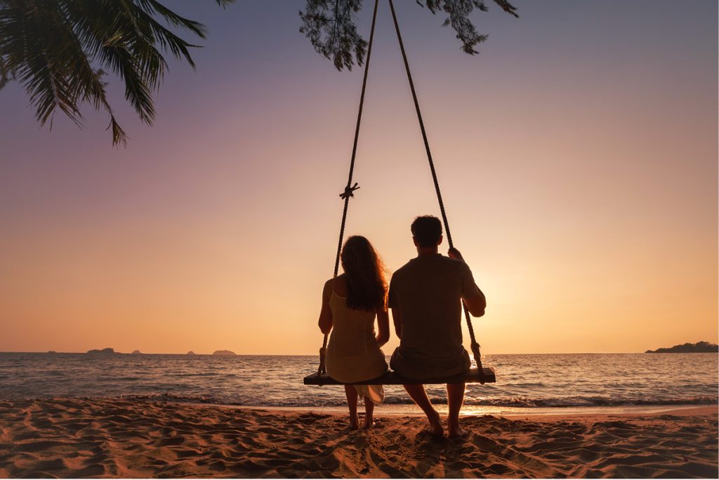 Couple sitting closely together on a large swing on a Bali beach, watching the sun set over the calm ocean horizon, symbolising connection and peaceful reflection.