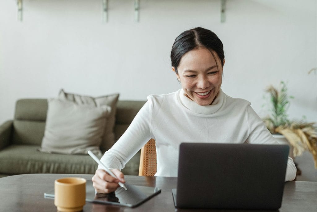 Female practitioner smiling at her computer while taking notes on her iPad, participating in online therapy or EFT training.